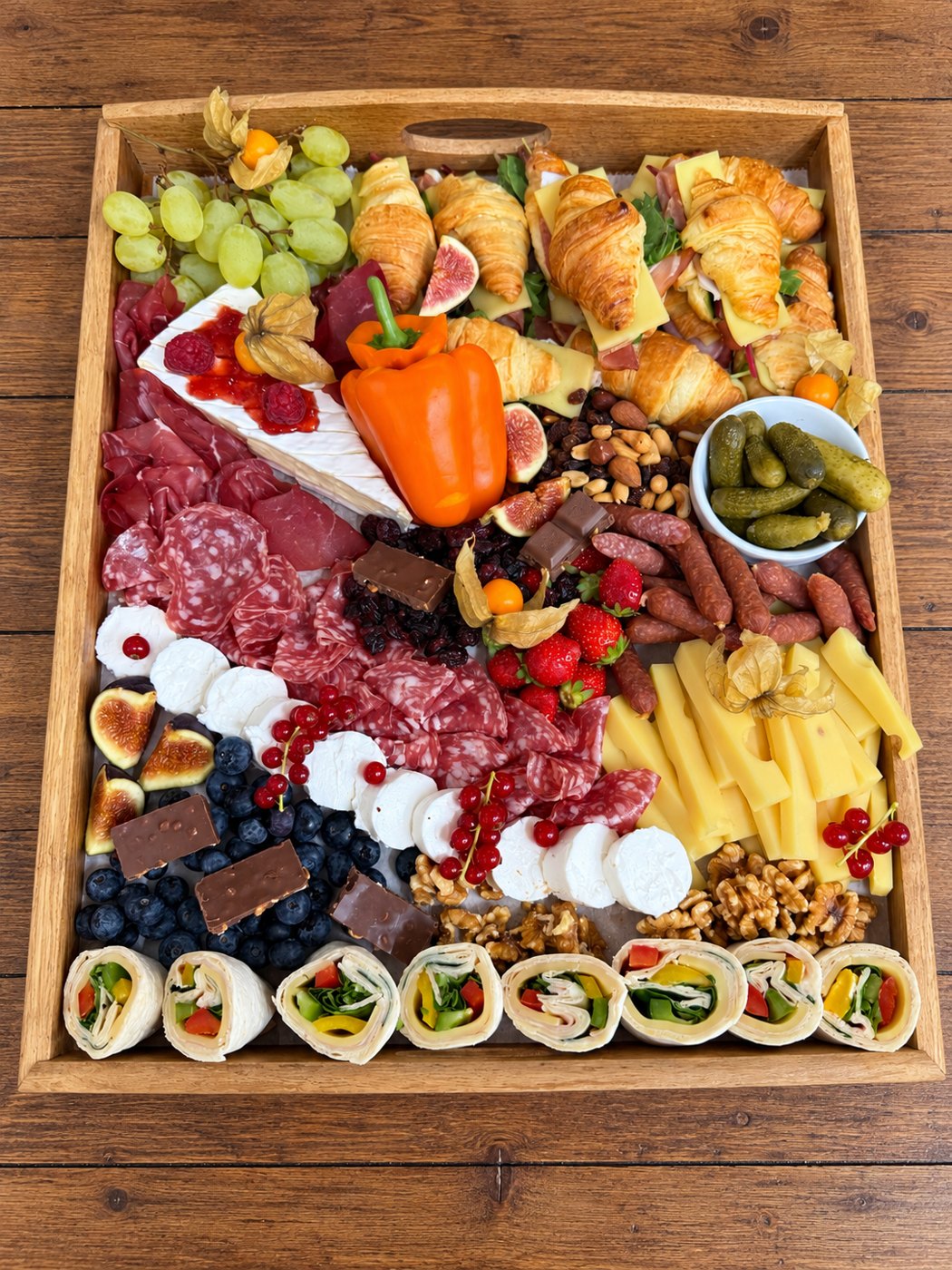 Platou mixt cu mini-croissante, brie, mezeluri, fructe, nuci și wrap-uri, pe masă de lemn